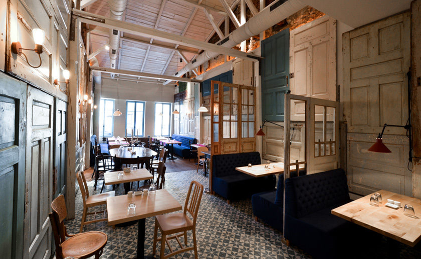 interior restaurante con mesas y sillas y bancos en terciopelo azul noche en capitoné. Puertas reutilizadas a modo de paneles colgadas en la pared