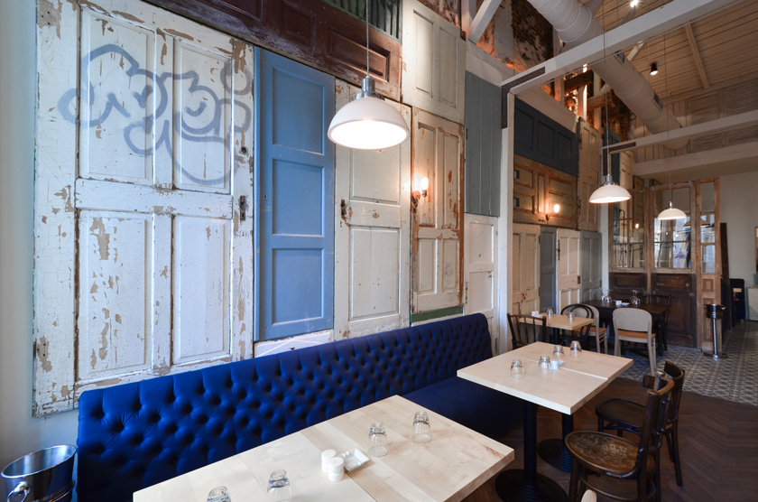 interior restaurante con banco alargado en terciopelo azul y capitoné. Mesas y sillas de madera. Puertas viejas colgadas en la pared a modo de panel.